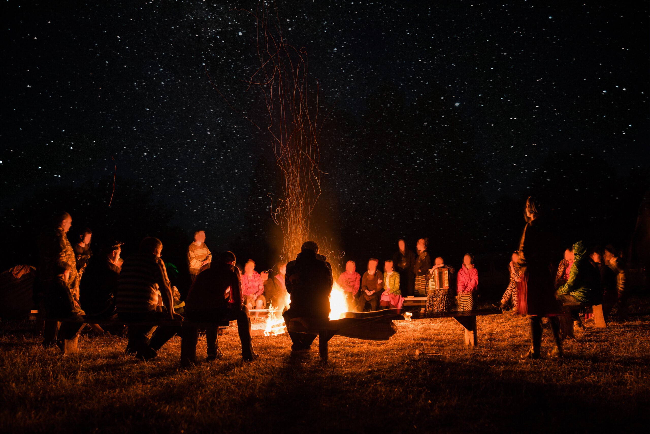 A large group of people stands around a blazing fire at night.