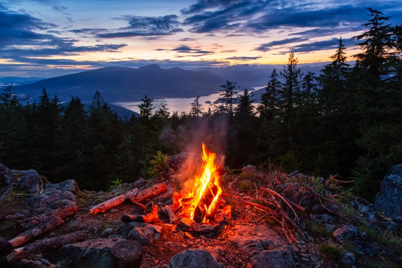 A campfire burns on top of a mountain at dusk overlooking a lake.