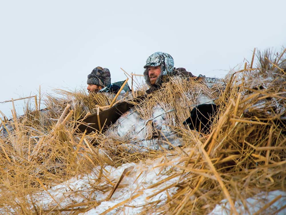 Two goose hunters sit in a snowy field. 