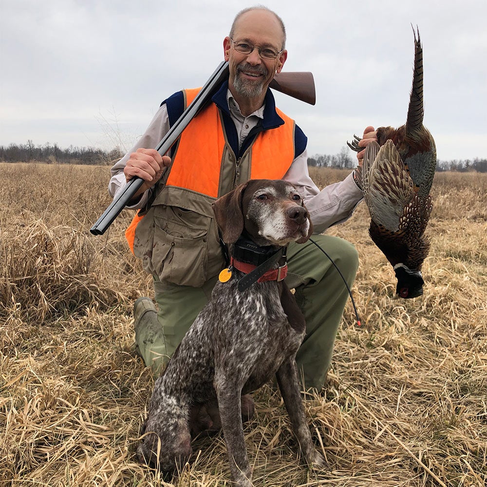 Phil Bourjaily kneeling next to hunting dog while holding a pheasant