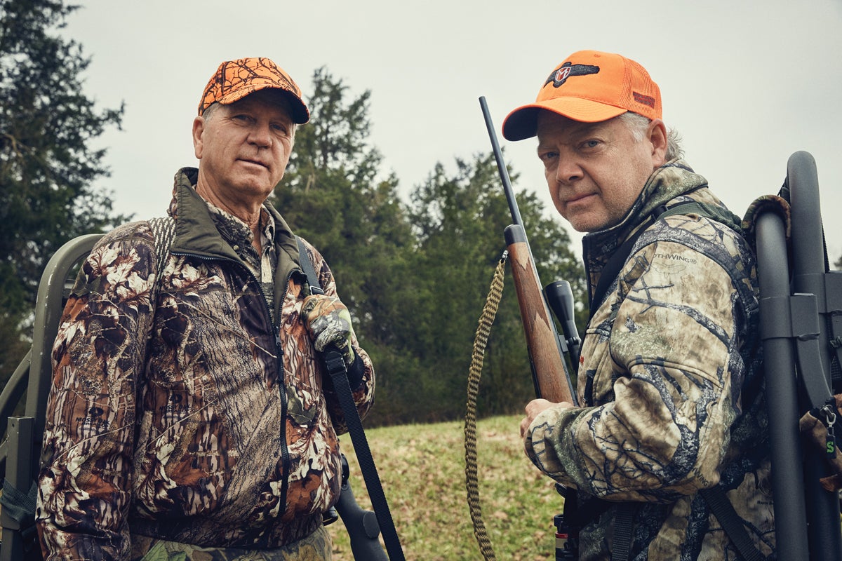 Two hunters stand in an open field holding rifles.