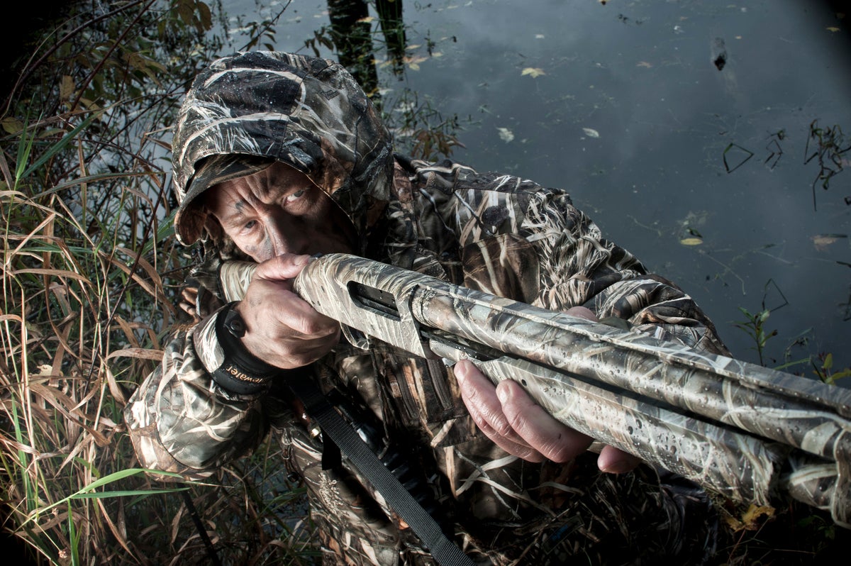 A hunter aiming a shotgun in the weeds.