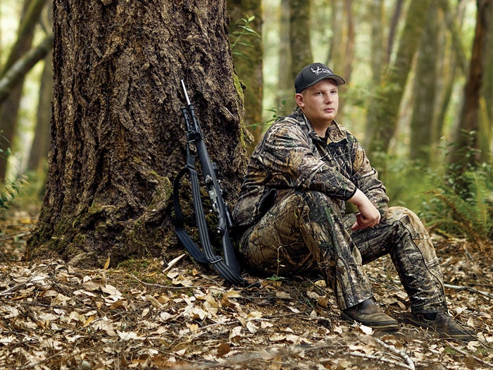 A young hunter in full camo gear sits by a tree in the woods.