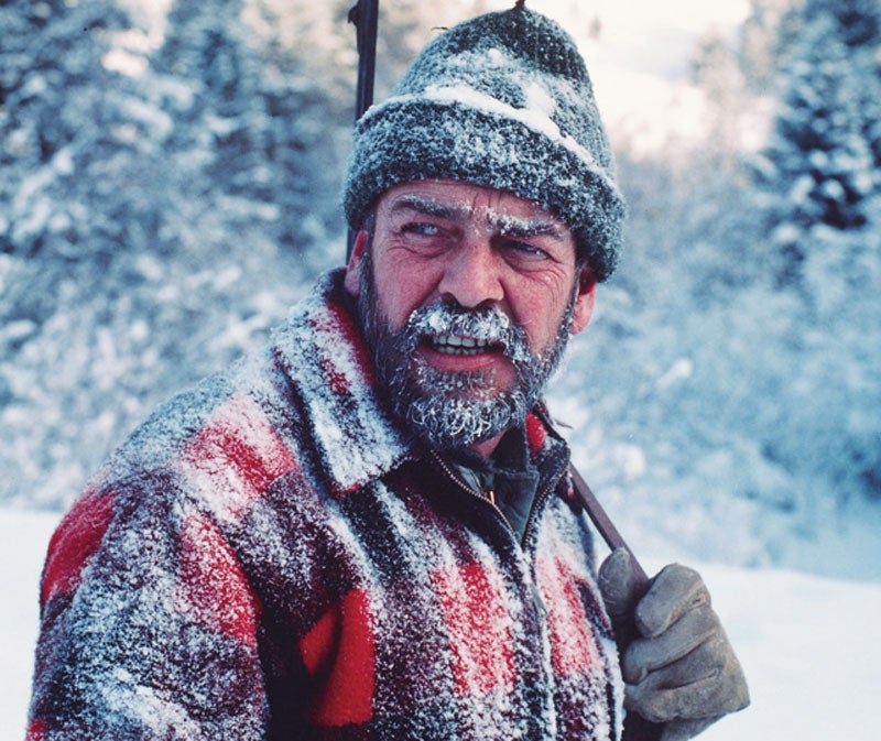 A hunter with a brown beard and snow frozen on his beard, while wearing a red plaid jacket.