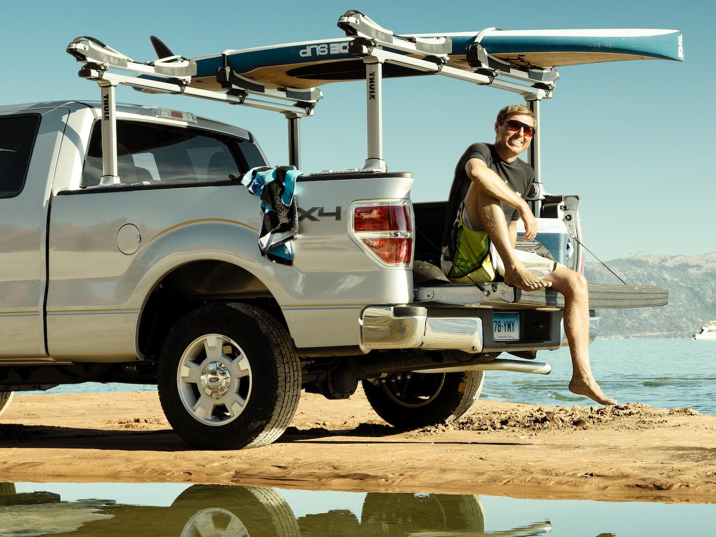 Guy sitting on the back of a pick-up truck with surfboard on rack.