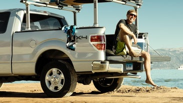 Guy sitting on the back of a pick-up truck with surfboard on rack.