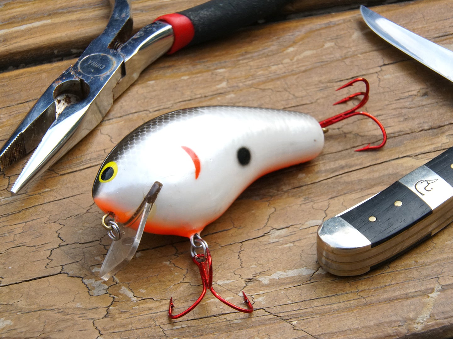 A crankbait, pliers, and knife on a wooden table.