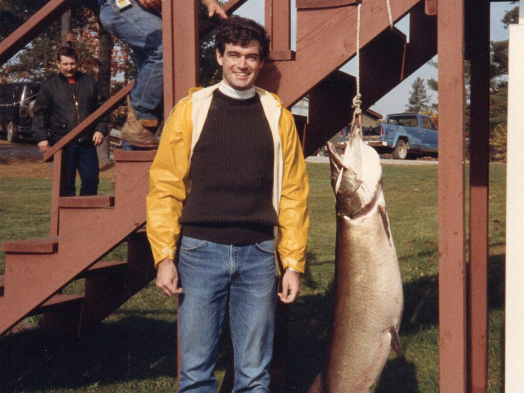 A young man stands beside a hanging muskie fish.