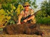 A hunter in camo, holding a winchester model 70 rifle, kneeling behind a feral hog.