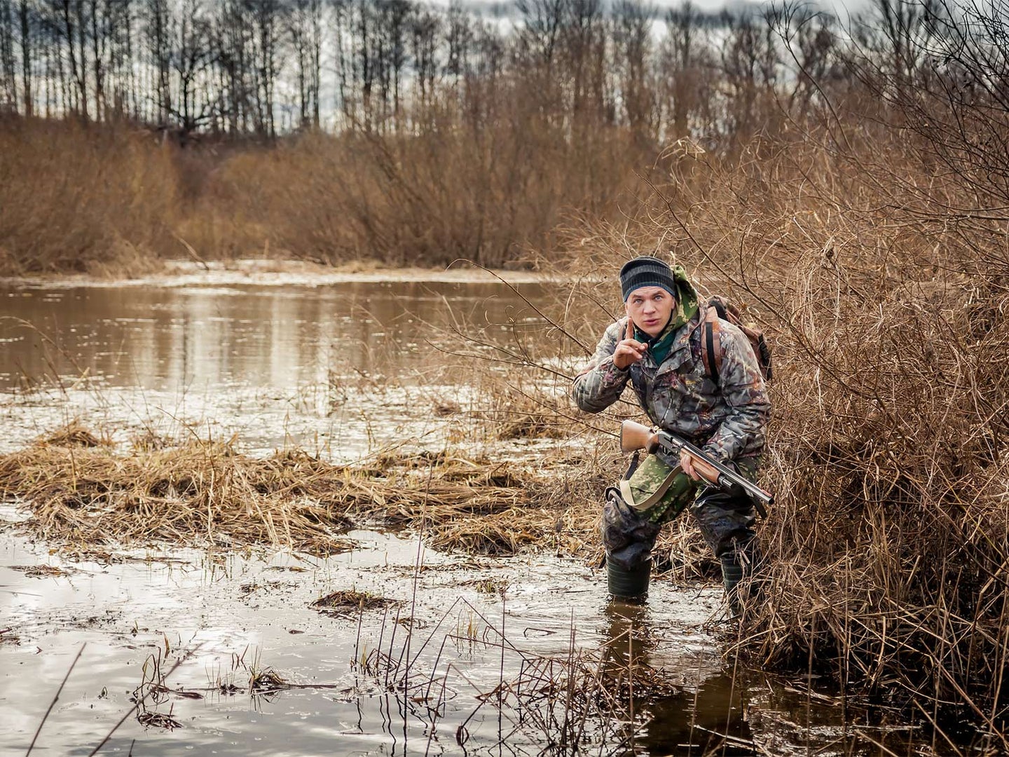 A hunter in camo stands near a brushline on a lake while holding a shotgun.