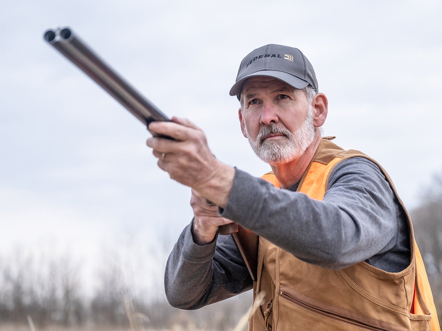 An older man in a vest and long sleeve shirt aims a shotgun in an open field.