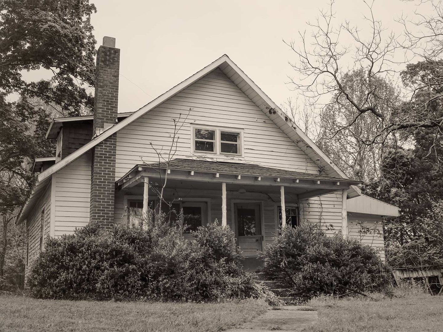 A black and white photo of an old farm house with peeling paint and oversized bushes growing beside the front porch.