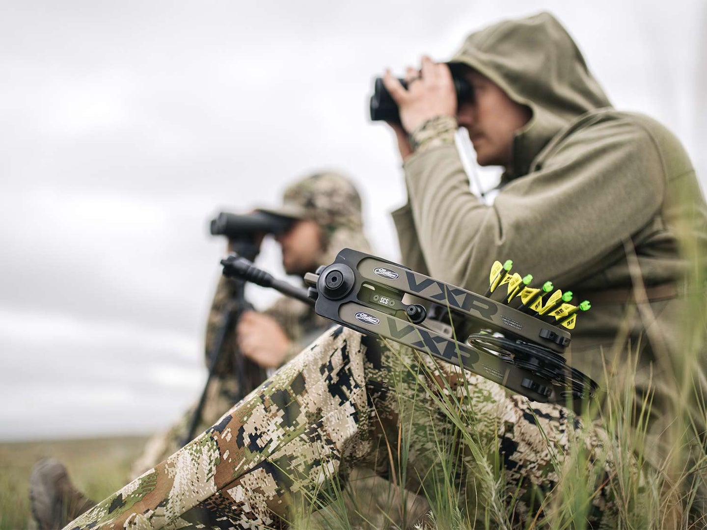 Two hunters scouting a mountainside through binoculars.