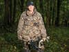 A woman hunter in full camo holds a compound bow and stands in the woods.