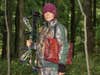 A female hunter in full camo holds a compound bow in the woods.