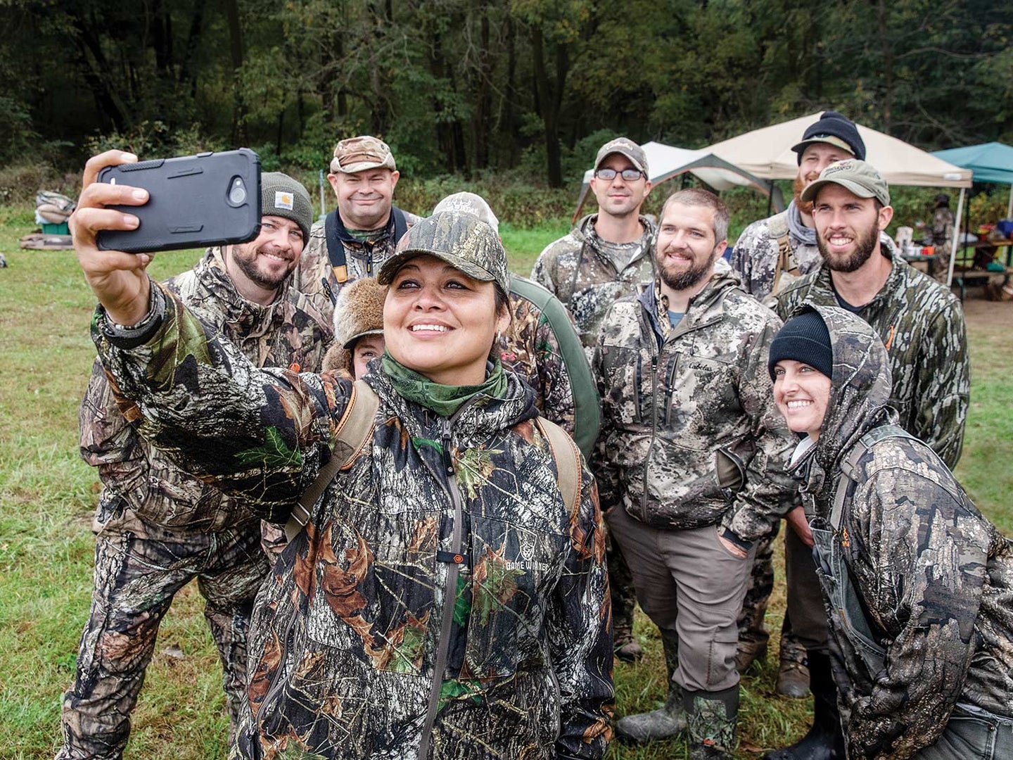 A group of men and women hunters in full camo pose for a selfie in an open field.