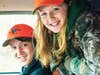 Two children, a bow and a girl, face the camera and smile while wearing hunting gear.