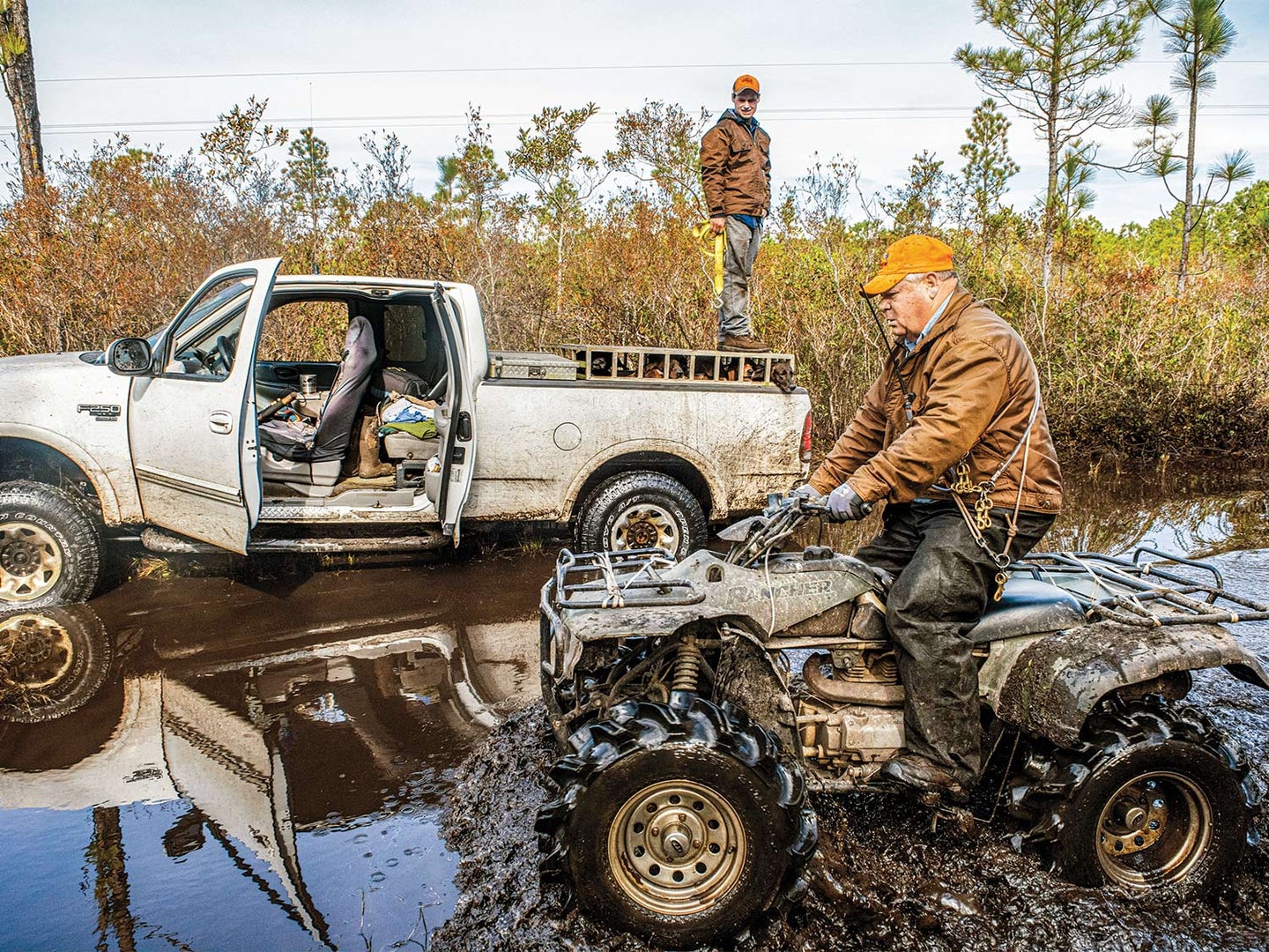 A hunter drives a four-wheeled ATV through the muddy water while one stands on top of a dog cage in the bed of a truck in the muddy water.