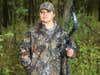 A woman hunter in a camo outfit holds a compound bow and stands in the woods.