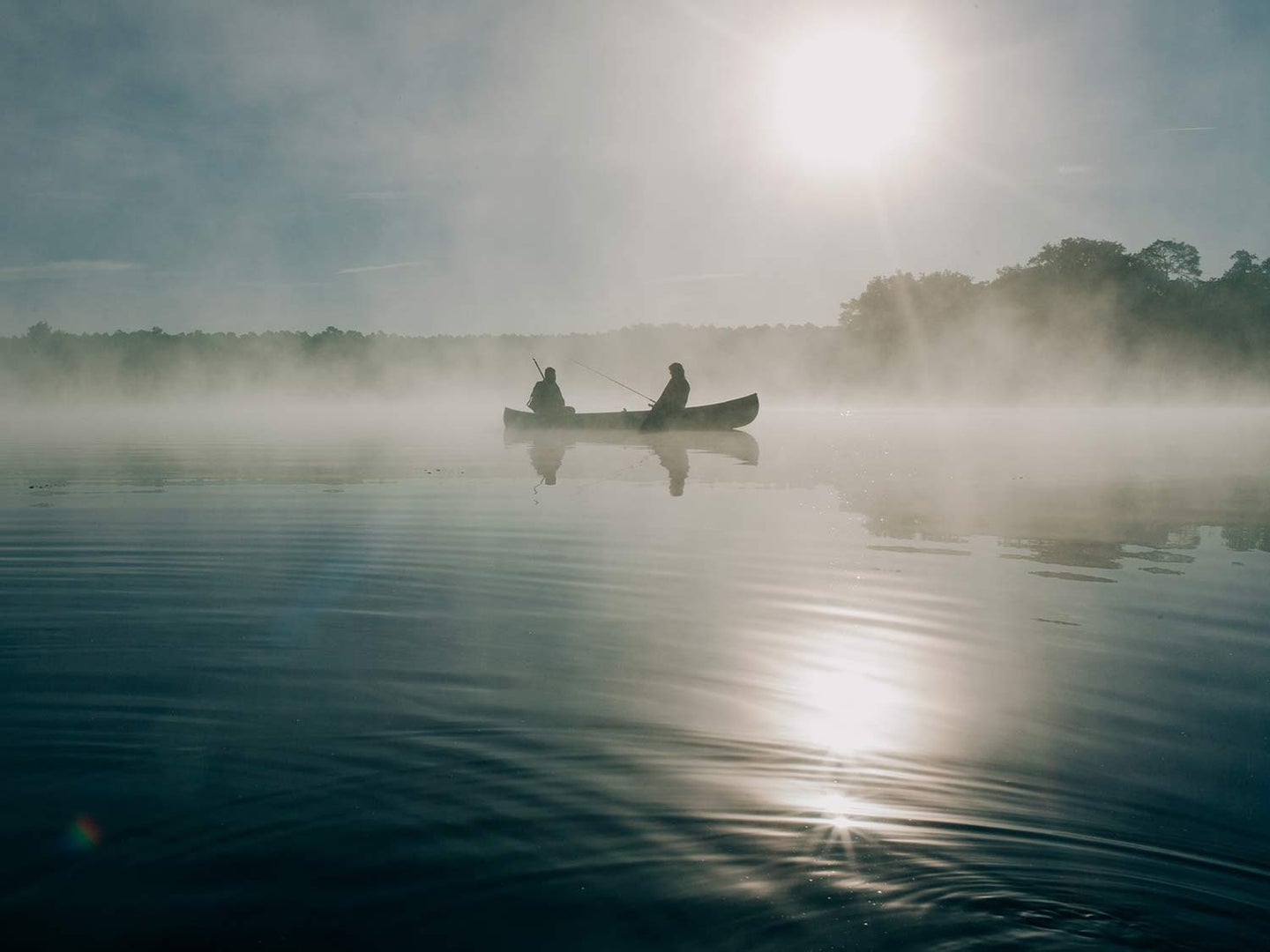 Two anglers sit and fish on a canoe in the middle of a morning sunrise, with fog rising off the smooth surface of the lake.