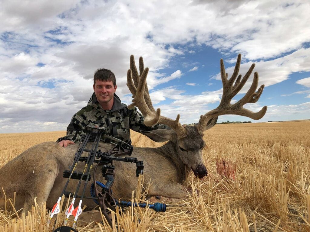 A hunter kneels behind a dead mule deer and holds up its head by the antlers.