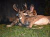 A woman hunter in full camo sits behind a dead whitetail buck at night.
