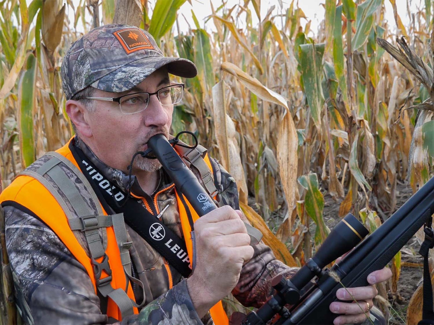 A hunter in camo and orange vest holds a rifle in one hand while blowing into a deer hunting grunt call held in the other.