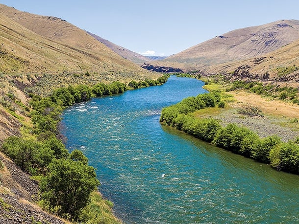 The lower Deschutes River flows through central Oregon. Lush greenery and rolling hills lines its banks.