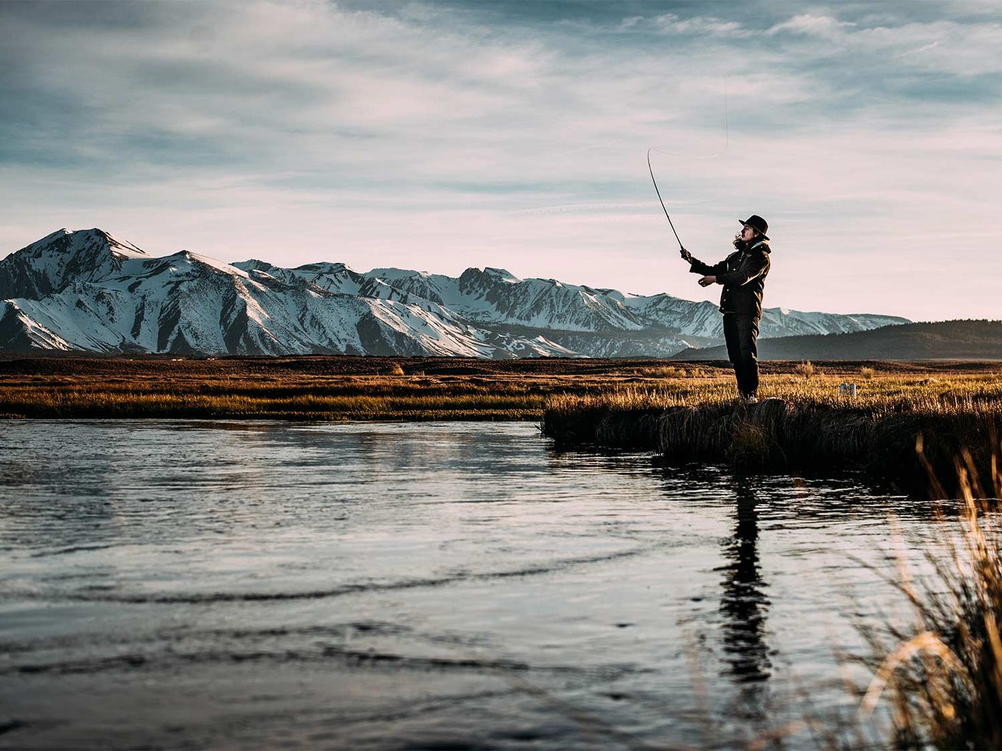 An angler fishing a river side with snowy mountains in the distance.