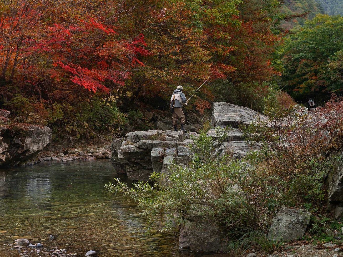 An angler stands on a rocky outcrop over a stream.