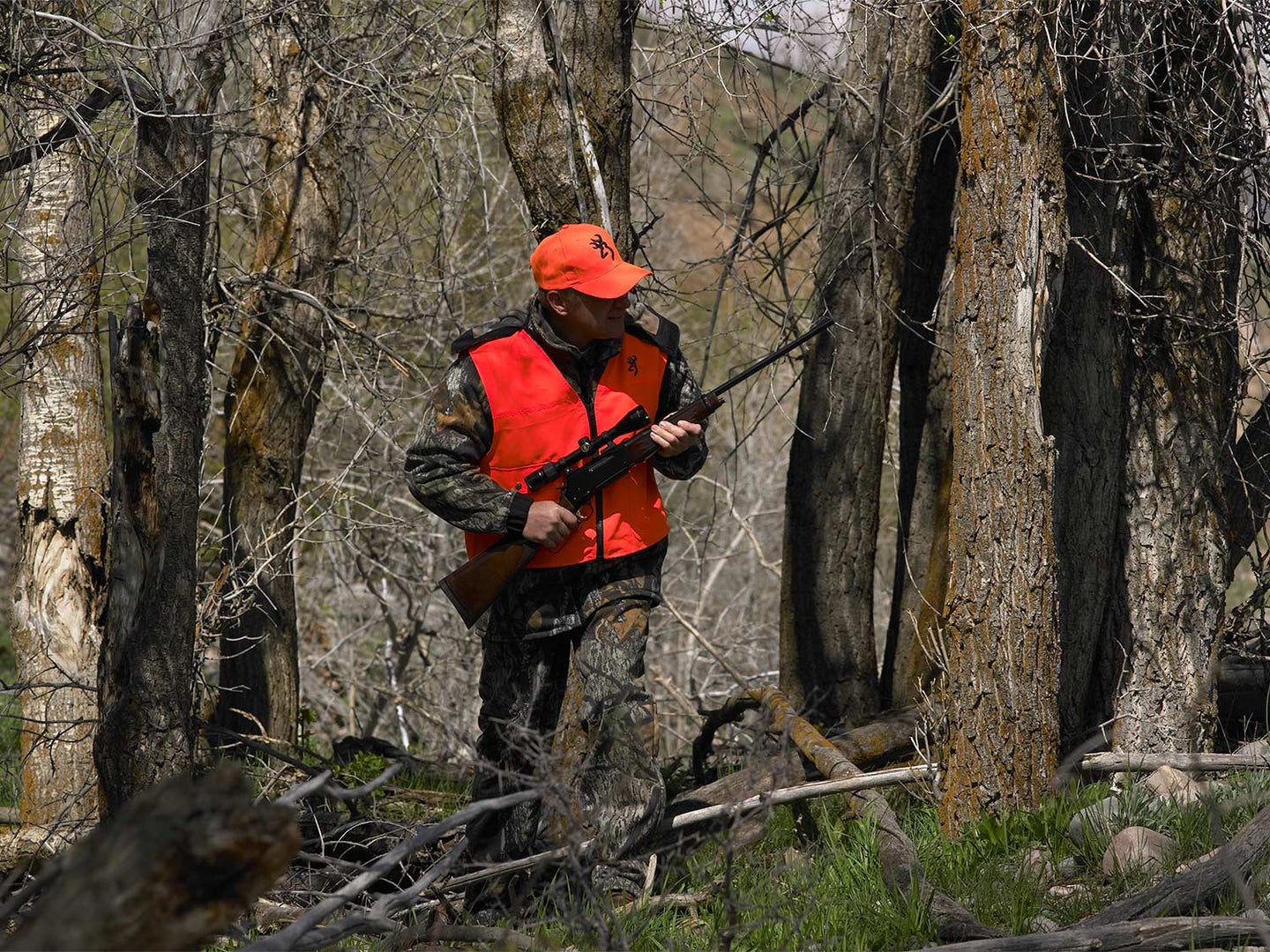 A hunter in camo and orange holding a rifle in the woods.