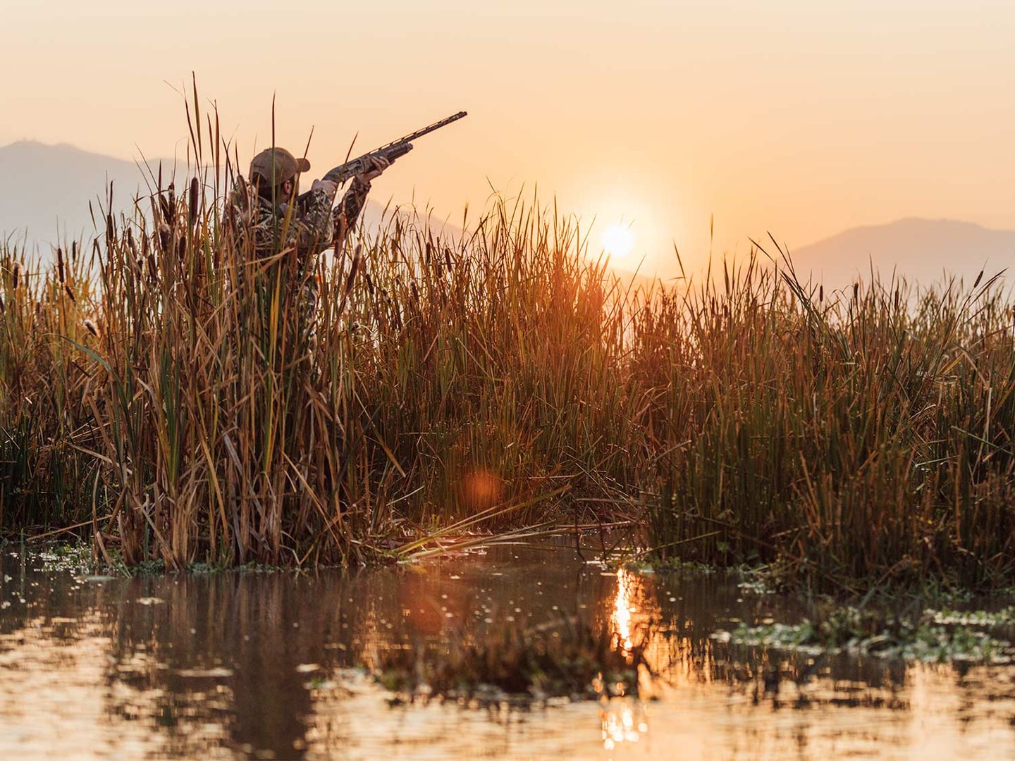 A waterfowl hunter stands in a marsh and aims a shotgun.