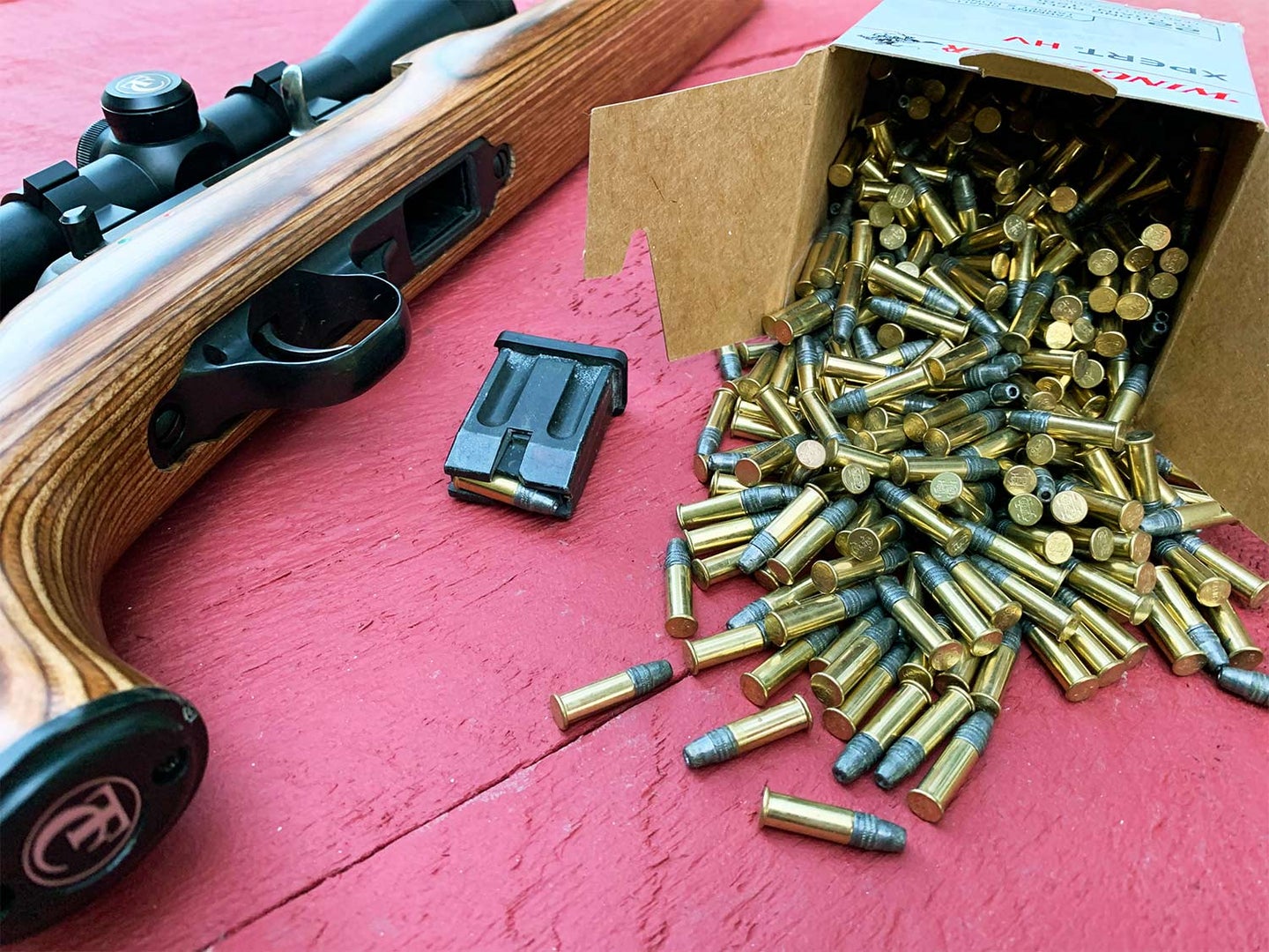 A rifle on a table next to a box of rifle ammunition spilling out onto the table.