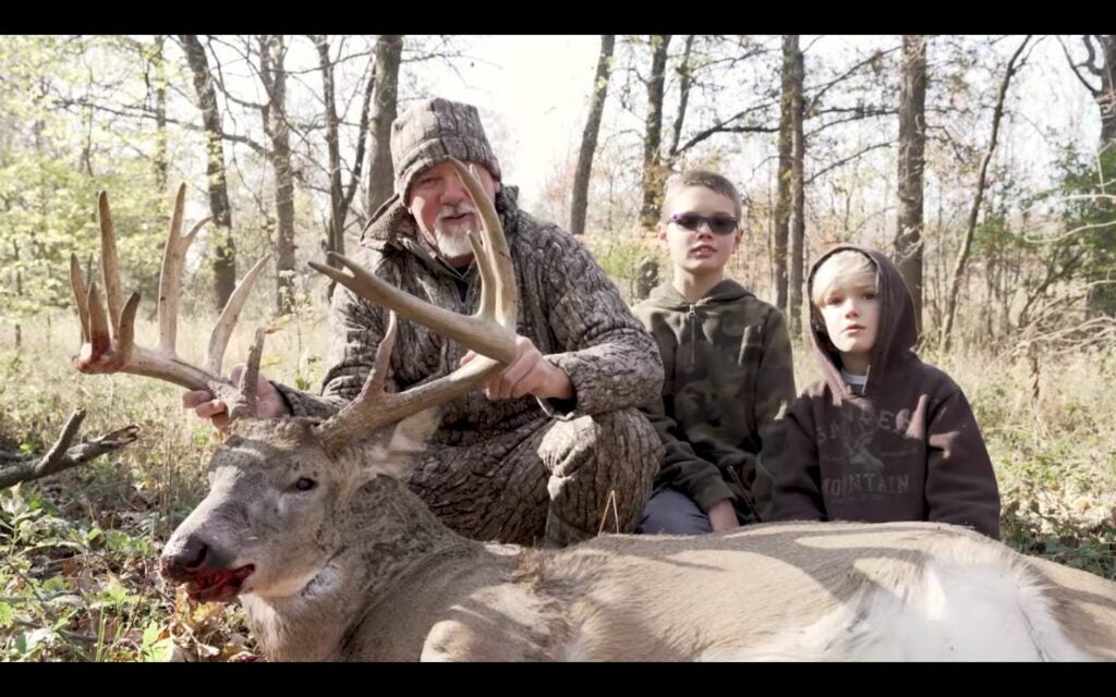 A hunter and two kids kneel behind a dropped whitetail deer.