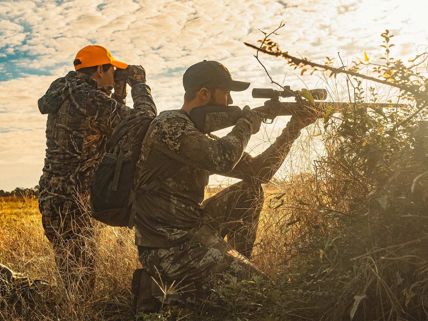 Two hunters kneel, scout, and aim their rifle in an open field.