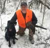 A hunter kneels in the snow and pets his hunting dog while holding a grouse.