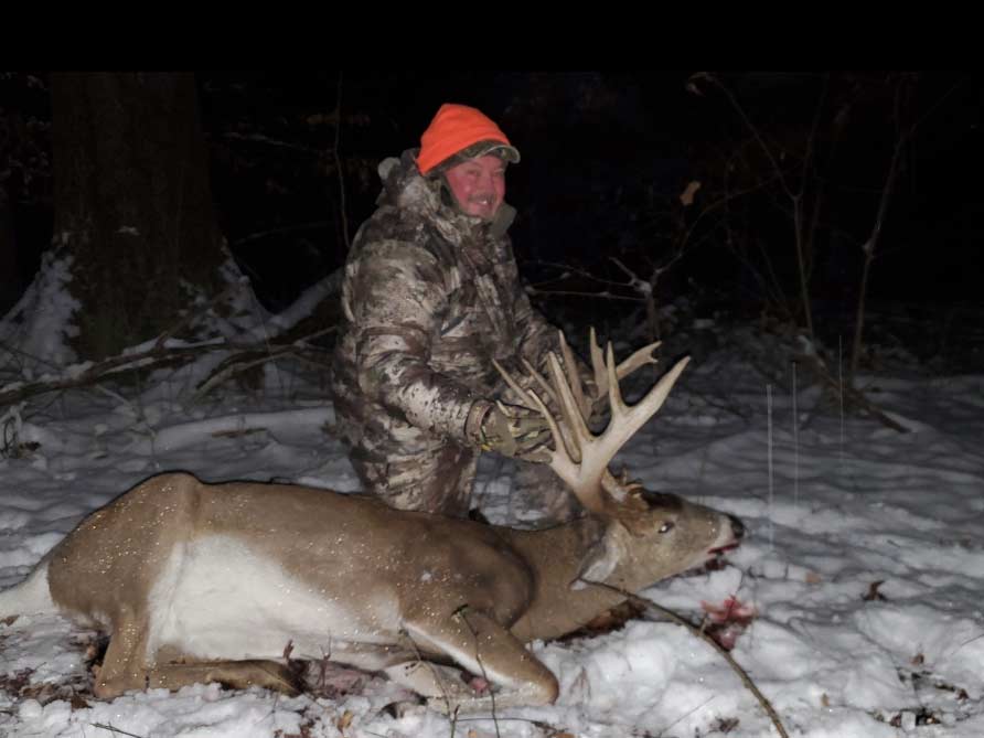 A hunter kneels in the snow next to a whitetail buck.