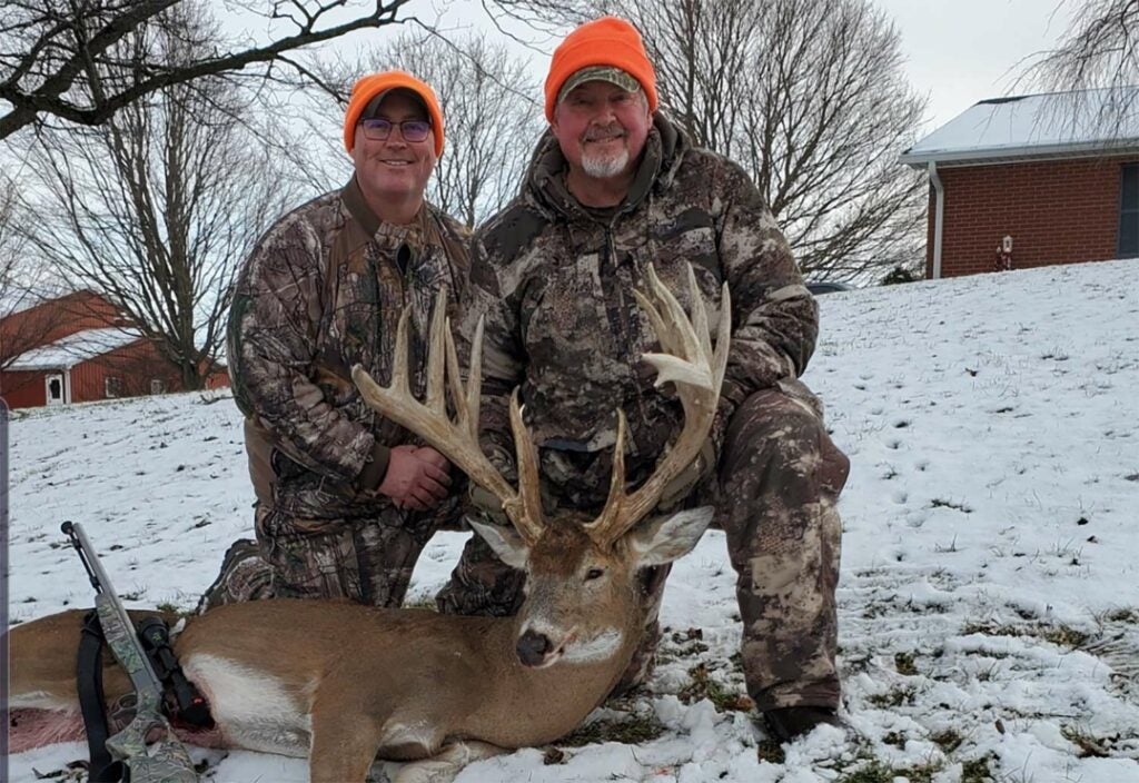 Two hunters next to a whitetail buck in the snow.
