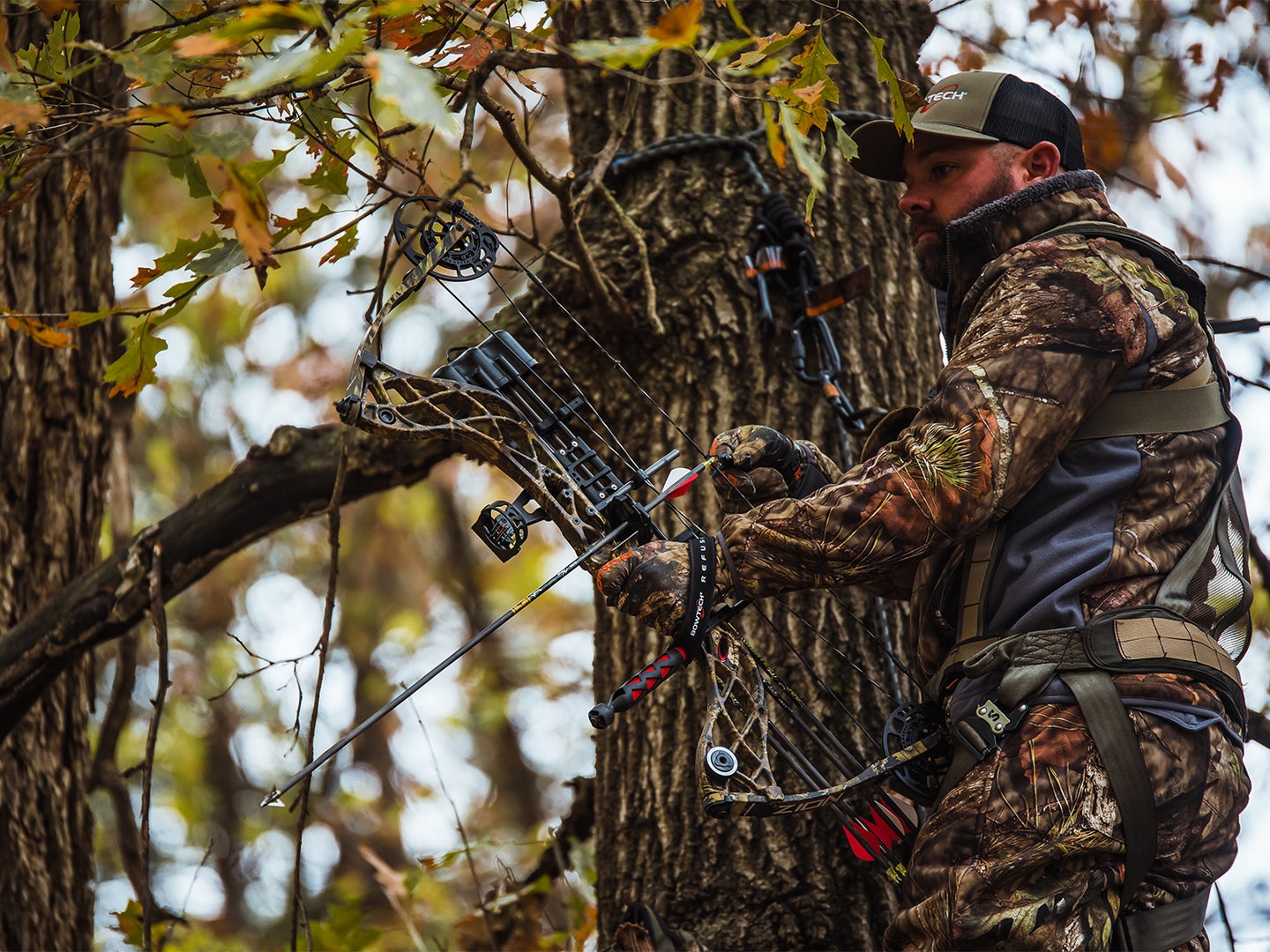A hunter stands in a treestand and holds a compound bow.
