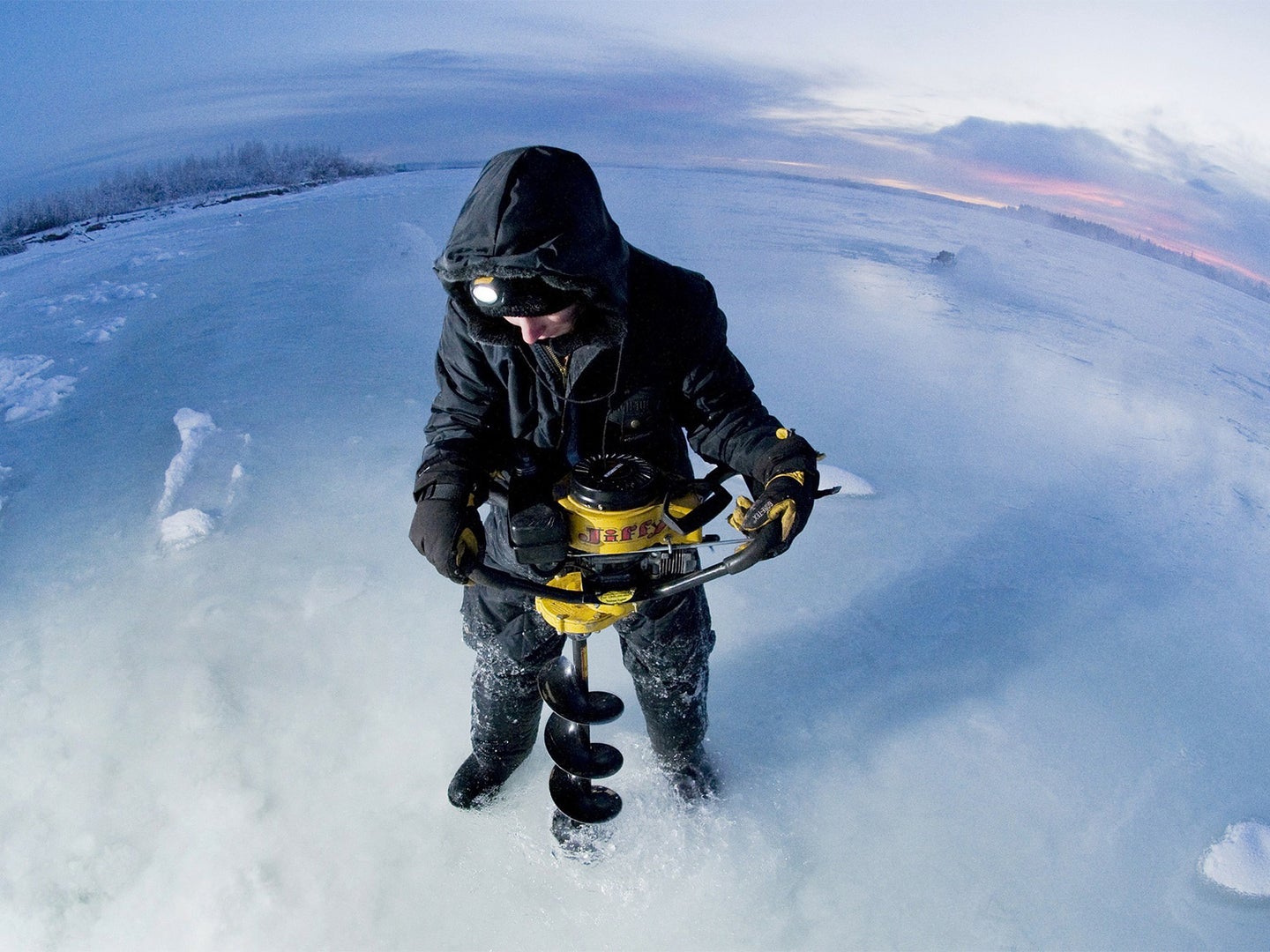 A man uses an ice auger to drill through the ice, while wearing the best heated jacket.