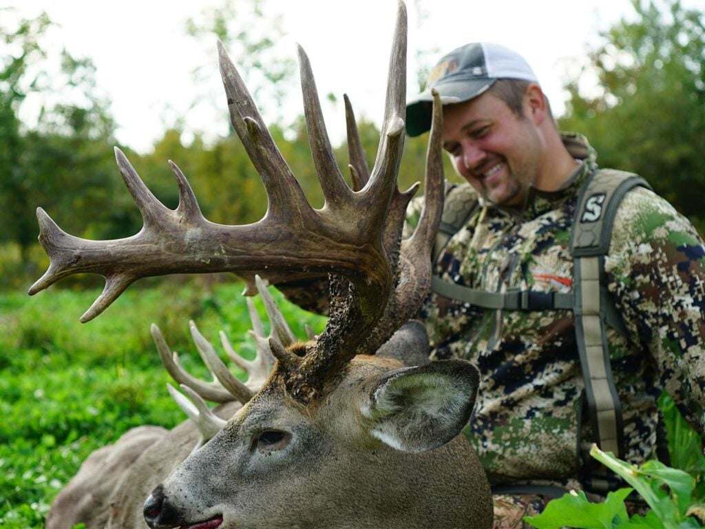 A hunter kneels beside a large whitetail deer and holds its head by the antlers.