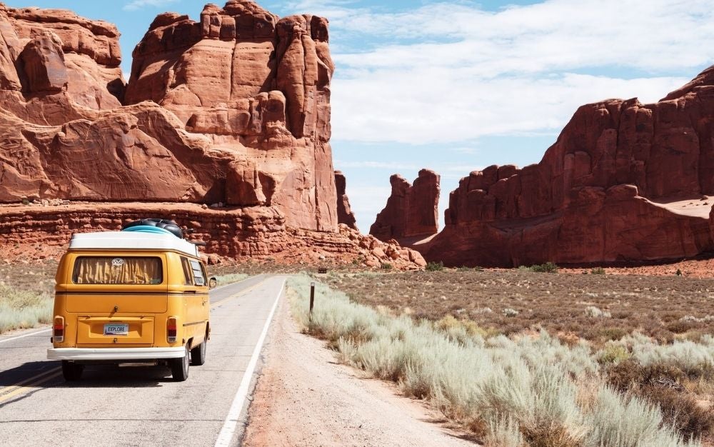 A yellow minibus traveling on open road between rocks and arches, sideways lush bushes.