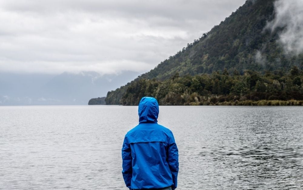 A man wearing a blue rainy jacket and standing in front of the big lake and under it the mountain.
