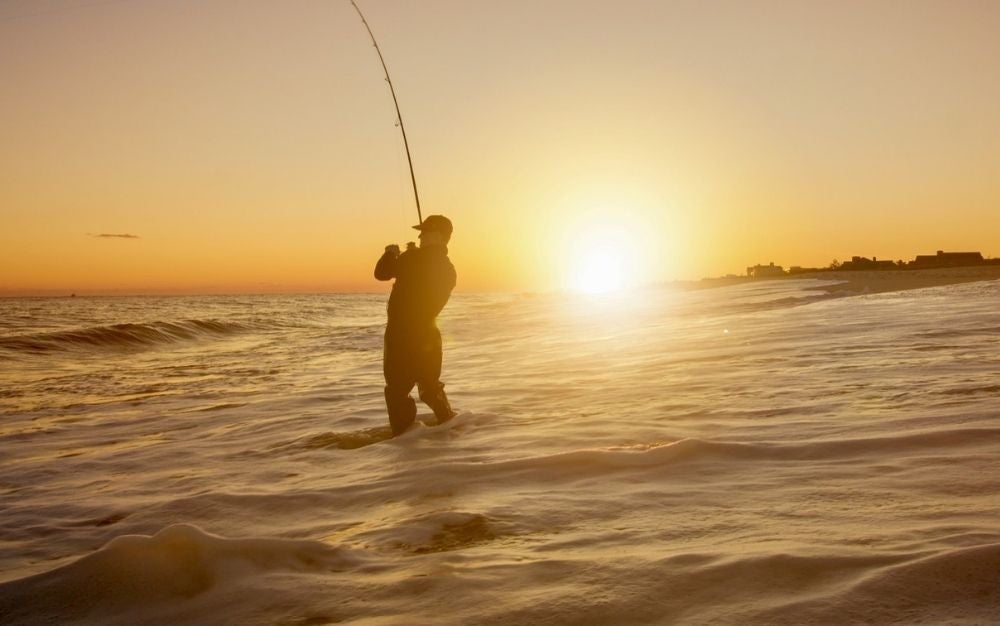 A man into the sea under the sunset with a fishing rolling reel in his hands fishing.