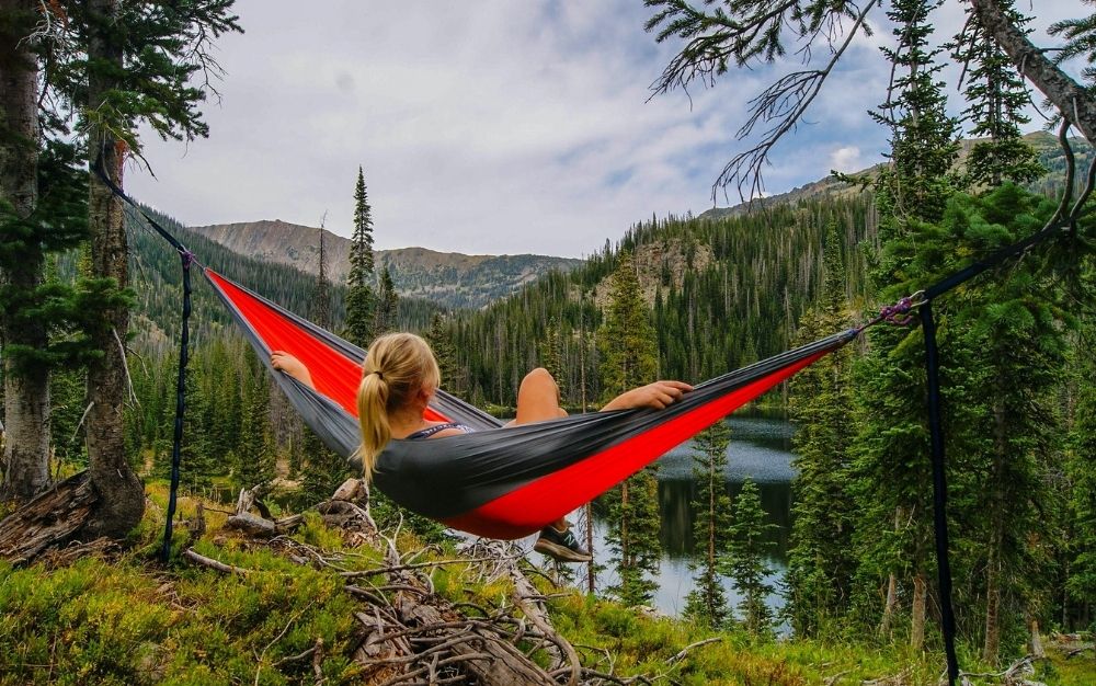 A woman with a view of mountain and a lake and relaxing over a black and red hammock.