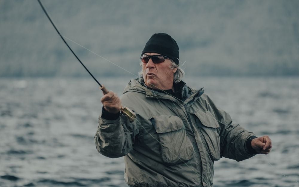 A man wearing a grey jacket, a black hat and black sunglasses in the middle of the water with a fishing stick fishing.