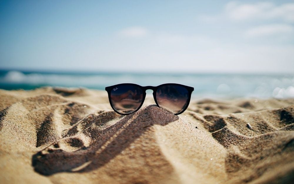 A pair of Ray Ban black sunglasses laying into the sand of the sea.