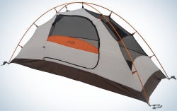 A green camping tent with orange and blue parts in it, as well as two ropes that are attached to the orange tent from above.