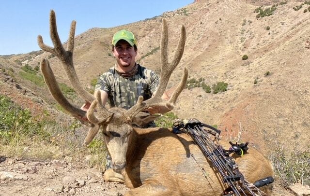 A boy grip and grins with a big mule deer and a bow in the desert