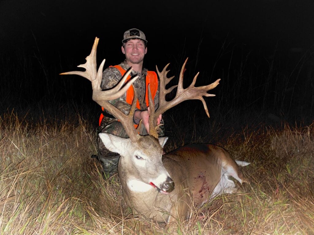 man in orange hunting vest poses behind dead deer with large rack at night